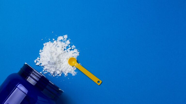 Cucharada de suplemento en polvo de creatina al lado de un recipiente azul sobre fondo azul intenso, ilustrando los esenciales de nutrición deportiva.