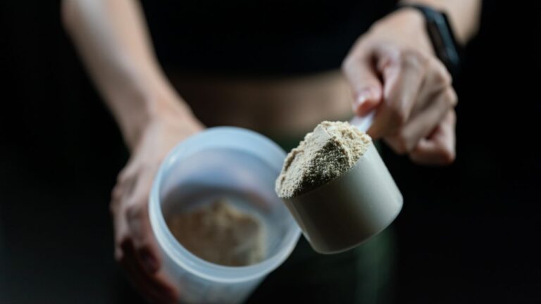 Mano sosteniendo medida de proteína en polvo con suplementos de fondo, resaltando la importancia de la nutrición pre y post entrenamiento.