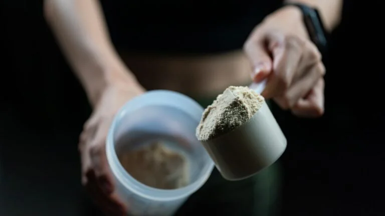 Mano sosteniendo medida de proteína en polvo con suplementos de fondo, resaltando la importancia de la nutrición pre y post entrenamiento.