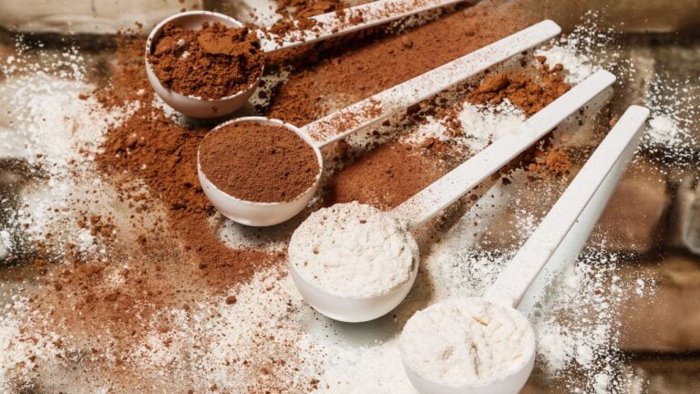 Cucharas con diferentes tipos de proteína en polvo y creatina sobre superficie de madera, relacionado con el aumento de masa muscular.