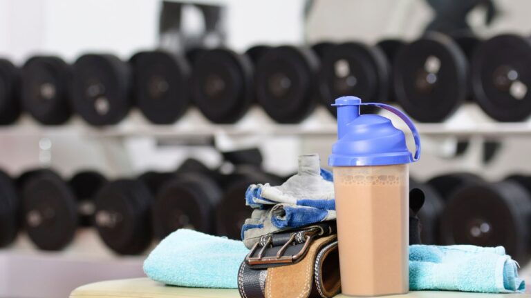 Batido de proteína listo junto a equipo de gimnasio, subrayando la comparativa en el uso de suplementos de creatina y proteína.