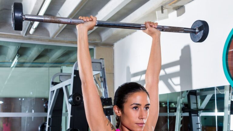Atleta completando una elevación de barra por encima de la cabeza en un gimnasio luminoso.