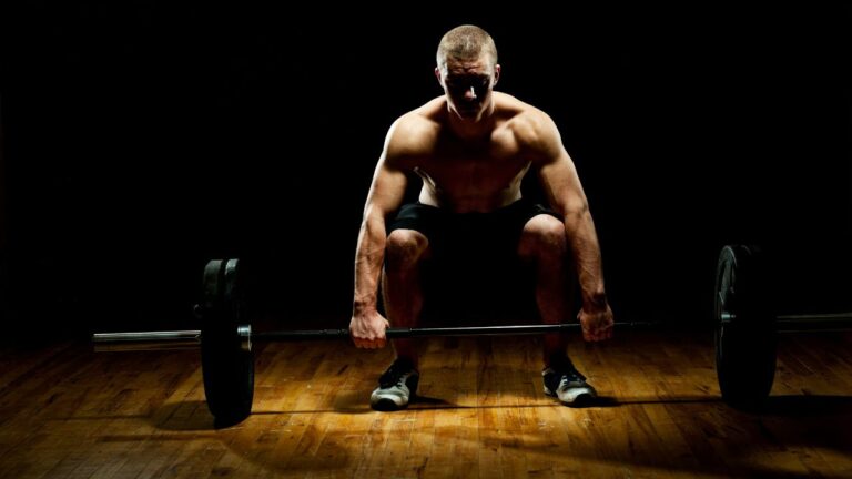Ejercicio de levantamiento de pesas en un entorno de gimnasio, iluminación focalizada en atleta y barra, destacando la técnica de ejecución.