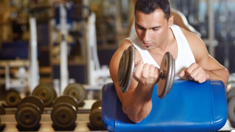 Hombre concentrado haciendo curl de bíceps con mancuernas en un gimnasio para fortalecer los músculos del brazo.