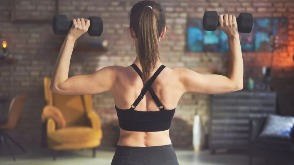Mujer entrenando hombros con mancuernas en casa, ilustrando una rutina de fortalecimiento de hombros para un entrenamiento doméstico.