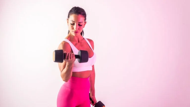Mujer realizando curls de bíceps con mancuerna en ambiente rosado, mostrando técnicas para fortalecer los brazos sin salir de casa.