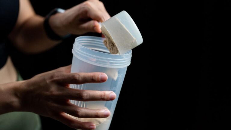 Manos sujetando un medidor lleno de polvo de proteína sobre un shaker, listo para mezclar antes de un entrenamiento.
