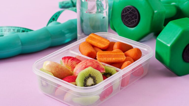 Táper con frutas variadas y pesas verdes sobre fondo rosa, combinación ideal para meriendas nutritivas en una rutina de definición muscular.