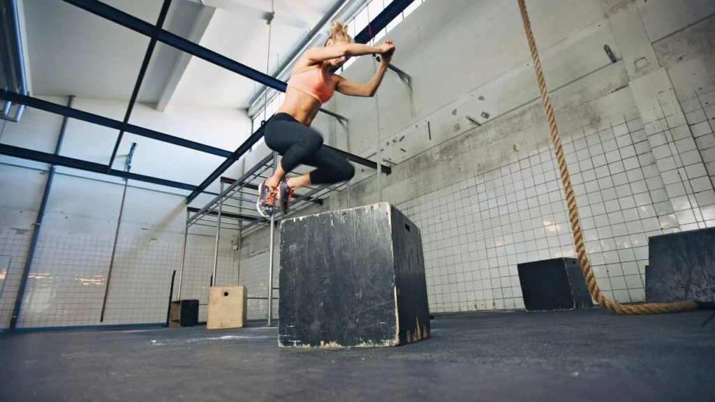 Una atleta salta sobre una caja de plyo en un gimnasio, un ejercicio que puede adaptarse a un WOD de CrossFit casero utilizando elementos comunes en casa.